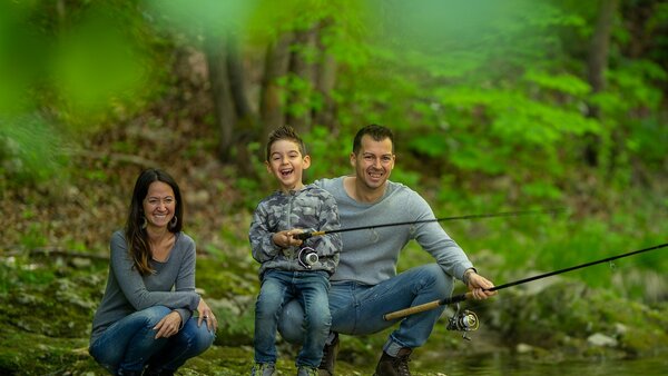 Eine Familie (Mann, Frau und Junge) sitzt unter Bäumen an einem Gewässer und angelt