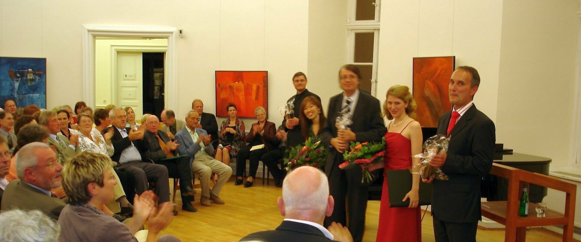 Fünf Musiker (drei Männer und zwei Frauen) stehen in einem mit Bildern geschmückten Raum vor einem Flügel und nehmen den Applas des vor ihnen sitzenden Publikums entgegen.