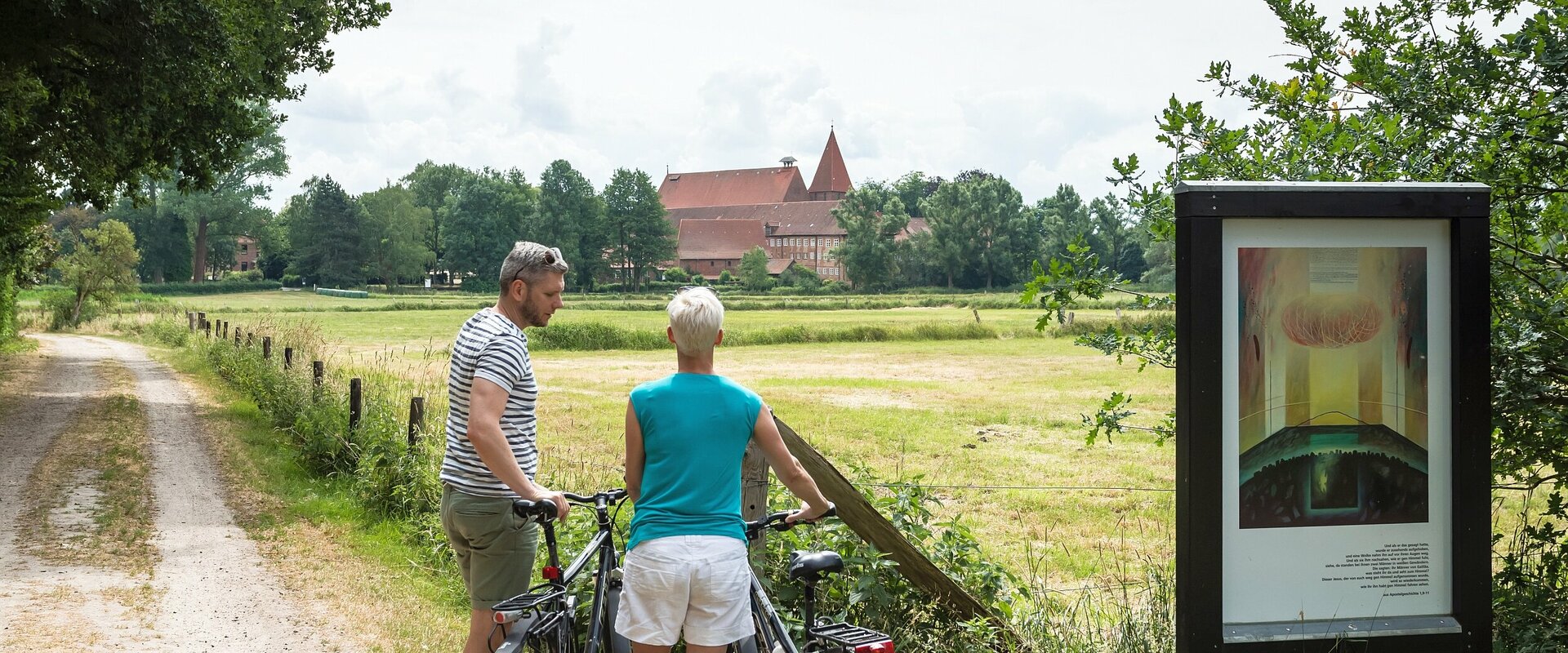 Ein sommerlich gekleidetes Paar steht mit seinen Fahrrädern auf einen Feldweg neben einer Tafel eines Besinnungsweges und blickt über Wiesen auf das entfernt liegende Kloster Ebstorf