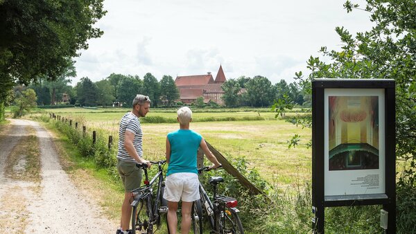 Ein sommerlich gekleidetes Paar steht mit seinen Fahrrädern auf einen Feldweg neben einer Tafel eines Besinnungsweges und blickt über Wiesen auf das entfernt liegende Kloster Ebstorf