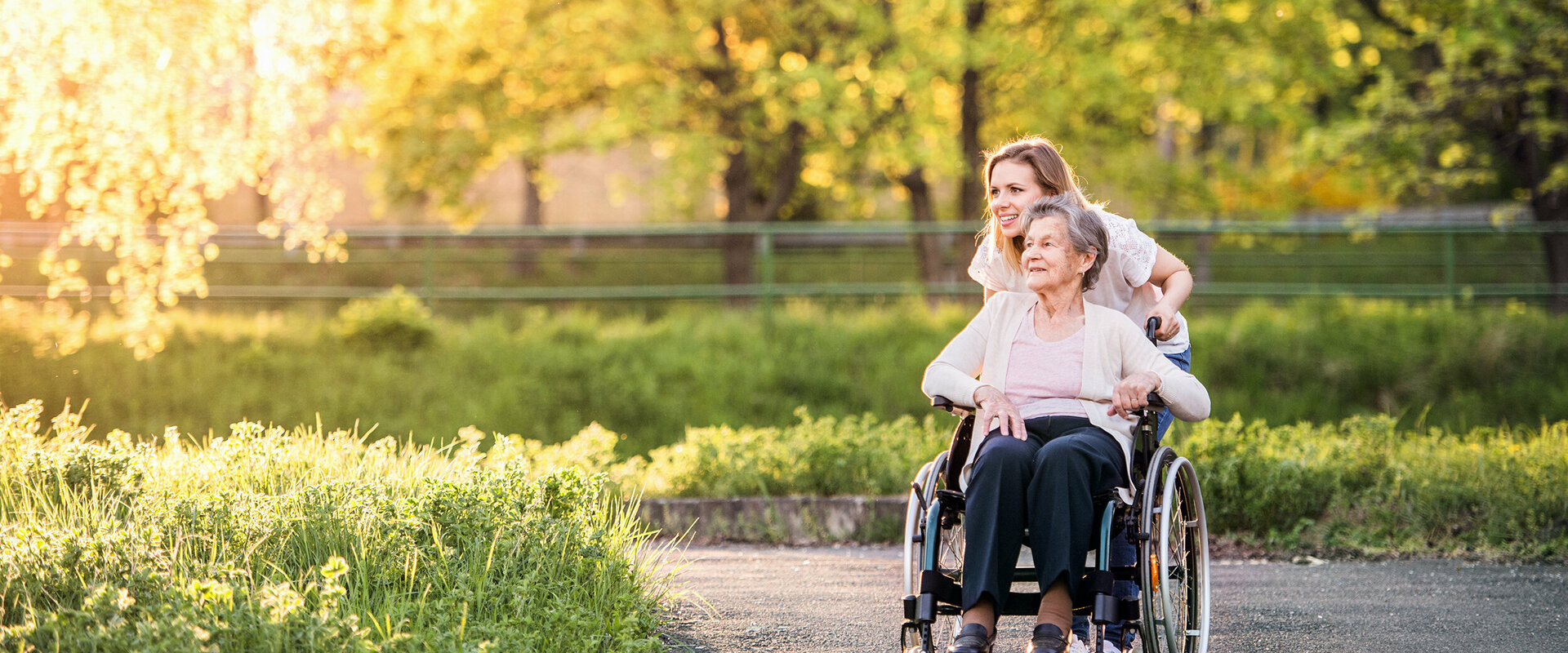 Seniorin / ältere Dame im Rollstuhl wird von einer jungen Frau auf einem sonnenbeschienenen, asphaltierten Weg geschoben. Links ein Blumenbeet, im Hintergrund Wiese, Blumen, Bäume und ein Zaun.