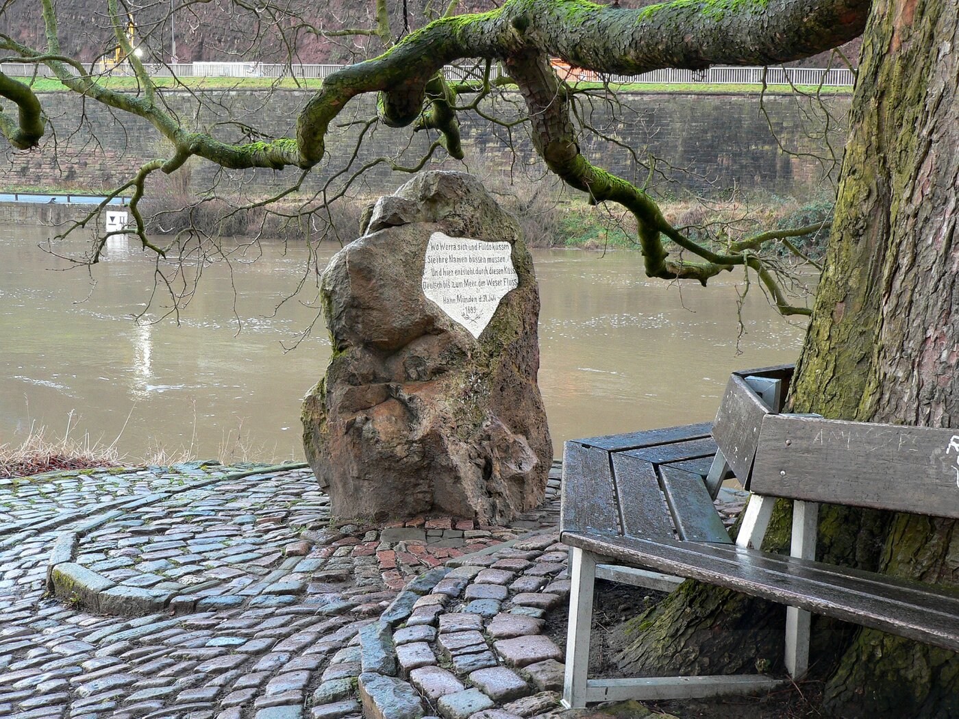 Der Alte Weserstein in Hannoversch Münden unter einem Baum mit Sitzbank vor der vorbeifließenden Weser.