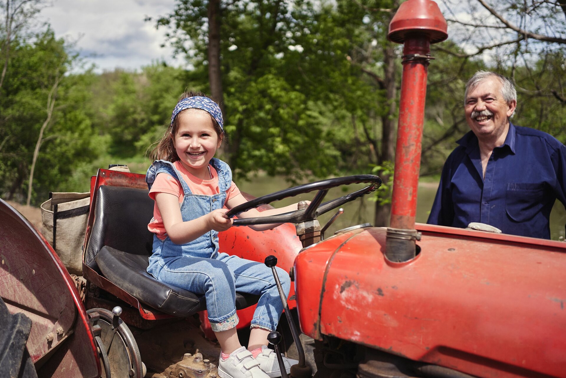 Ein kleines Mädchen sitzt lachend auf dem Sitz eines älteren roten Traktors. Hinter dem Traktor steht ein lachender älterer, grauhaariger Mann.