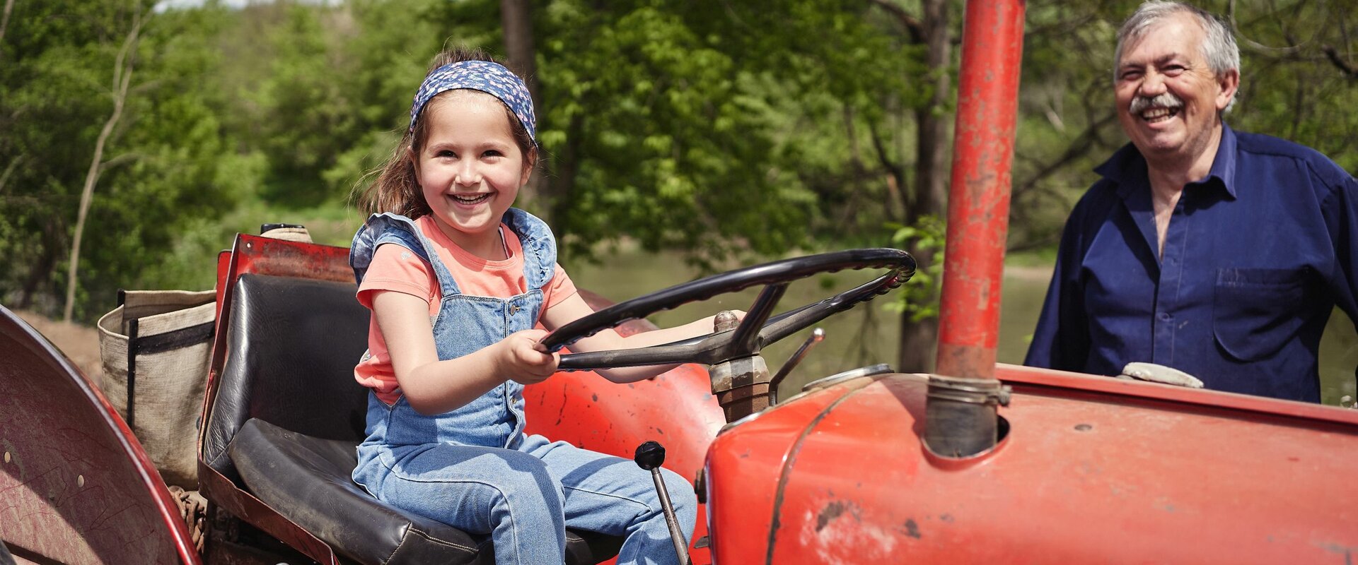 Ein kleines Mädchen sitzt lachend auf dem Sitz eines älteren roten Traktors. Hinter dem Traktor steht ein lachender älterer, grauhaariger Mann.