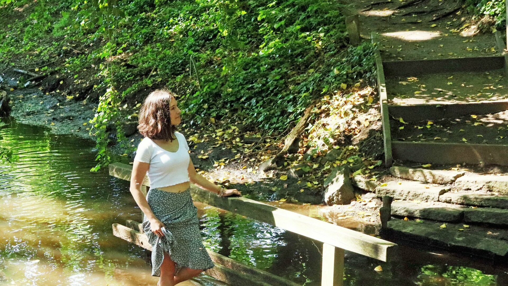 Eine junge Frau mit weißer Bluse und getupftem Rock steht barfuß im Wasser eines Baches, der zum 400-Wasser-Barfußpfad in Bad Bodenteich gehört.