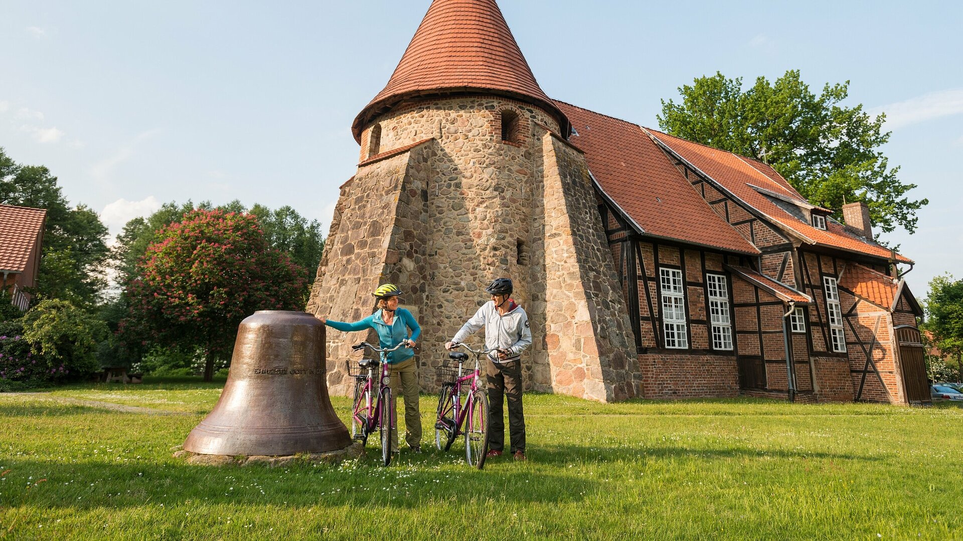 Ein Paar steht mit seinen Fahrrädern neben einer alten Kirchenglocke, die auf der Wiese vor der Radwegkirche St. Remigius in Suderburg liegt.