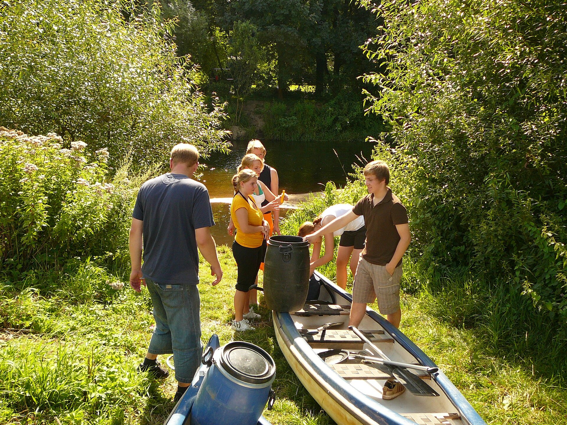 Eine Gruppe junger Männer und Frauen bereitet sich auf den Start zu einer Kanutour vor. Vor ihnen ein auf einer Wiese liegendes Kanu und mehrere Bootstonnen, hinter ihnen der Fluss