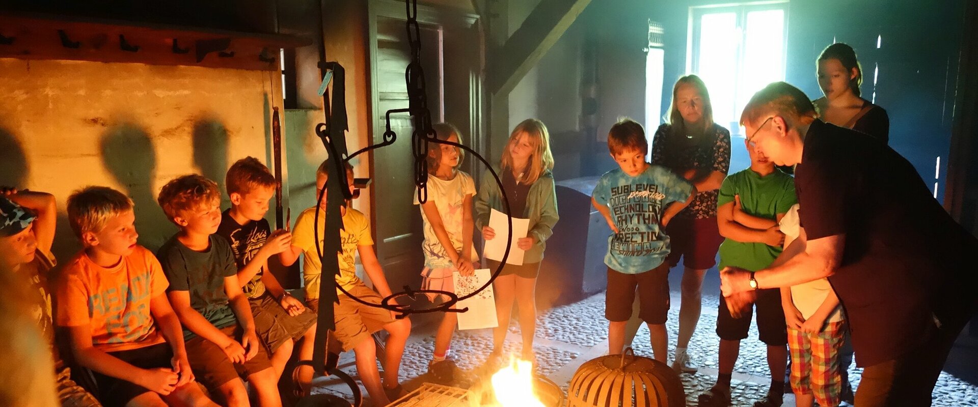 Eine Kindergruppe sitzt oder steht im Kreis um das brennende offene Feuer in einem Niedersachsenhaus und lauscht den Erklärungen einer Museumspädagogin