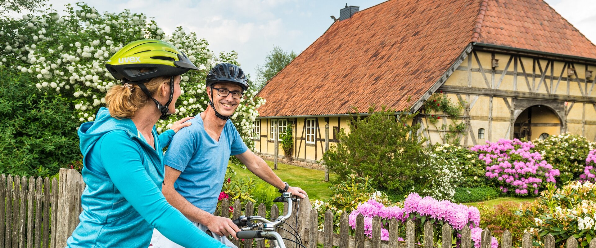 Ein Paar steht mit seinen Fahrrädern vor dem blühenden Vorgarten eines in Fachwerk gebauten Niedersachsenhauses.