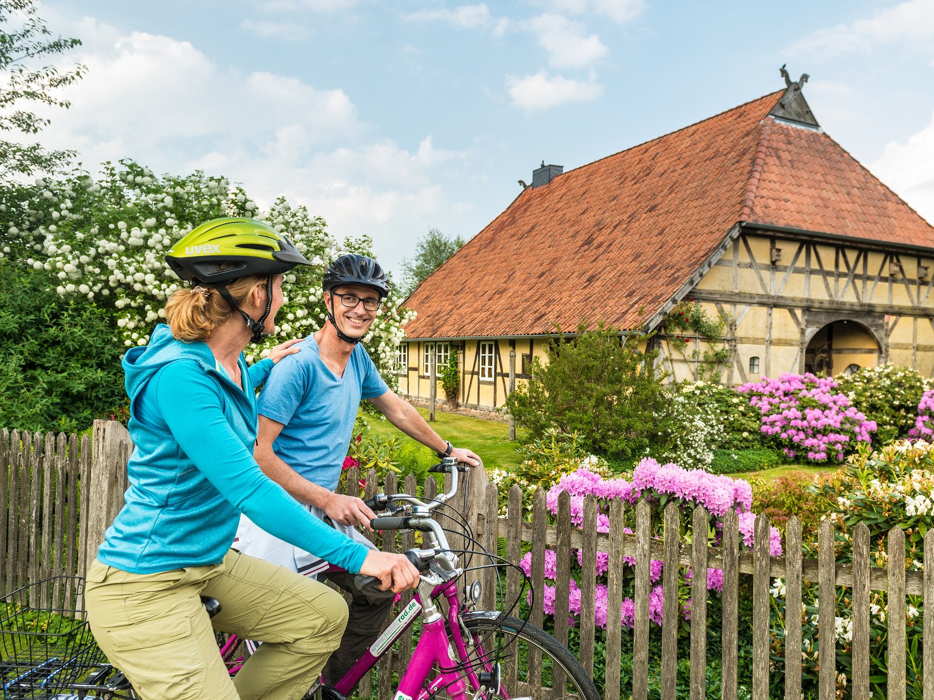 Ein Paar steht mit seinen Fahrrädern vor dem blühenden Vorgarten eines in Fachwerk gebauten Niedersachsenhauses.