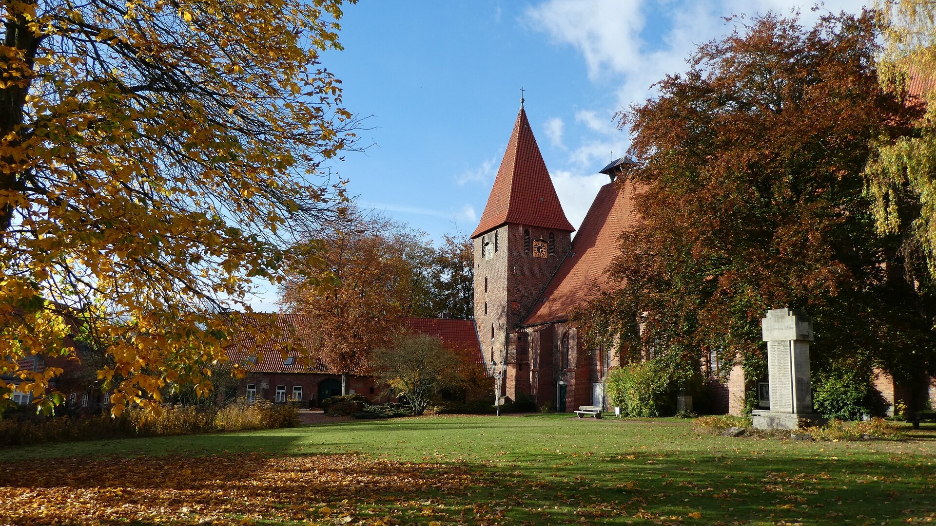 Bäume mit Herbstlaub, eine Wiese und blauer Himmel umrahmen den Kirchturm und die angrenzenden Gebäude des Klosters Ebstorfs