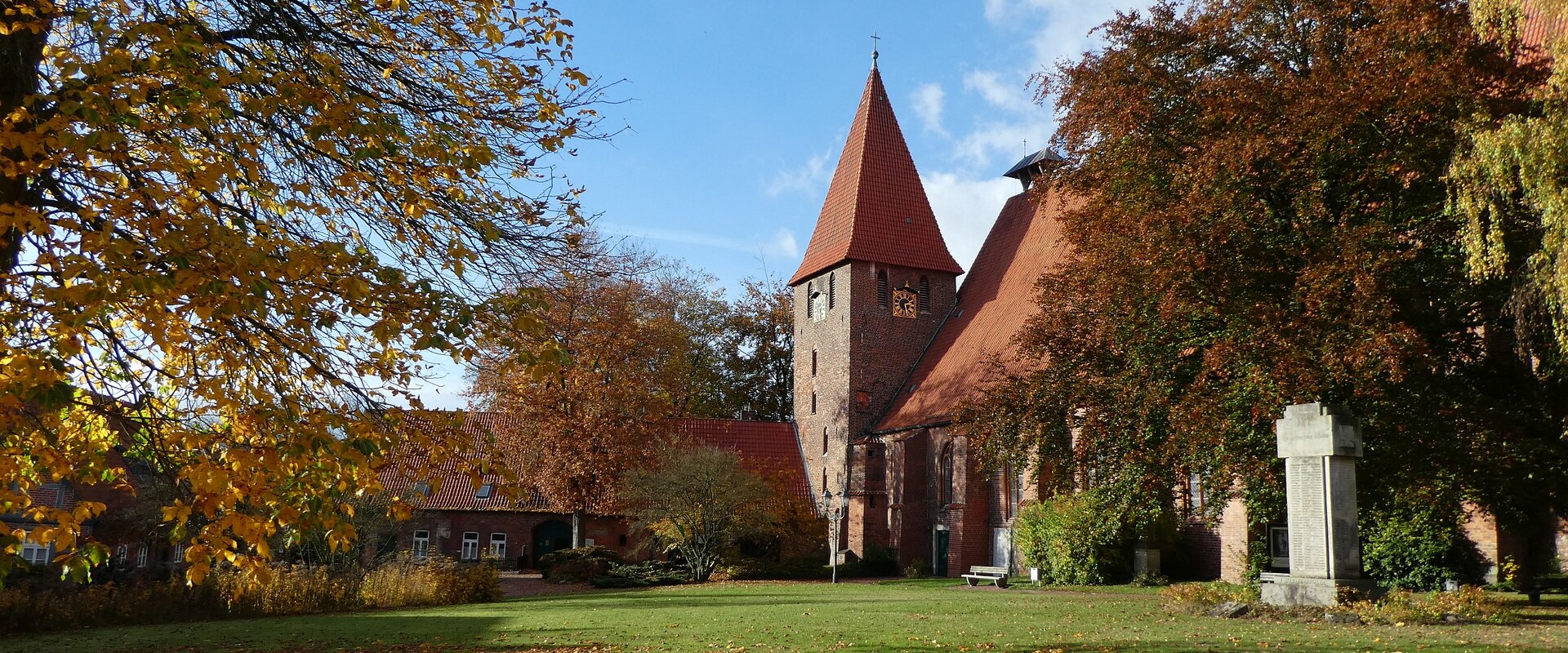 Bäume mit Herbstlaub, eine Wiese und blauer Himmel umrahmen den Kirchturm und die angrenzenden Gebäude des Klosters Ebstorfs