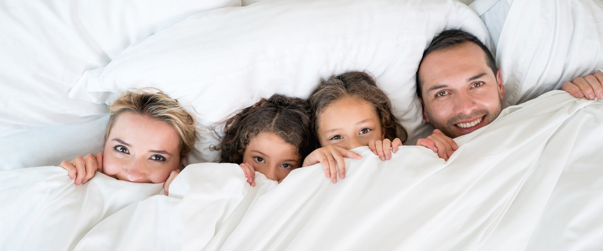 Eine Familie Frau, zwei Kinder und ein Mann, liegt nebeneinander in einem großen Bett. Die weiße Bettdecke haben sie mit den Händen bis unters Kinn gezogen, sodass nur die Köpfe hervorschauen.