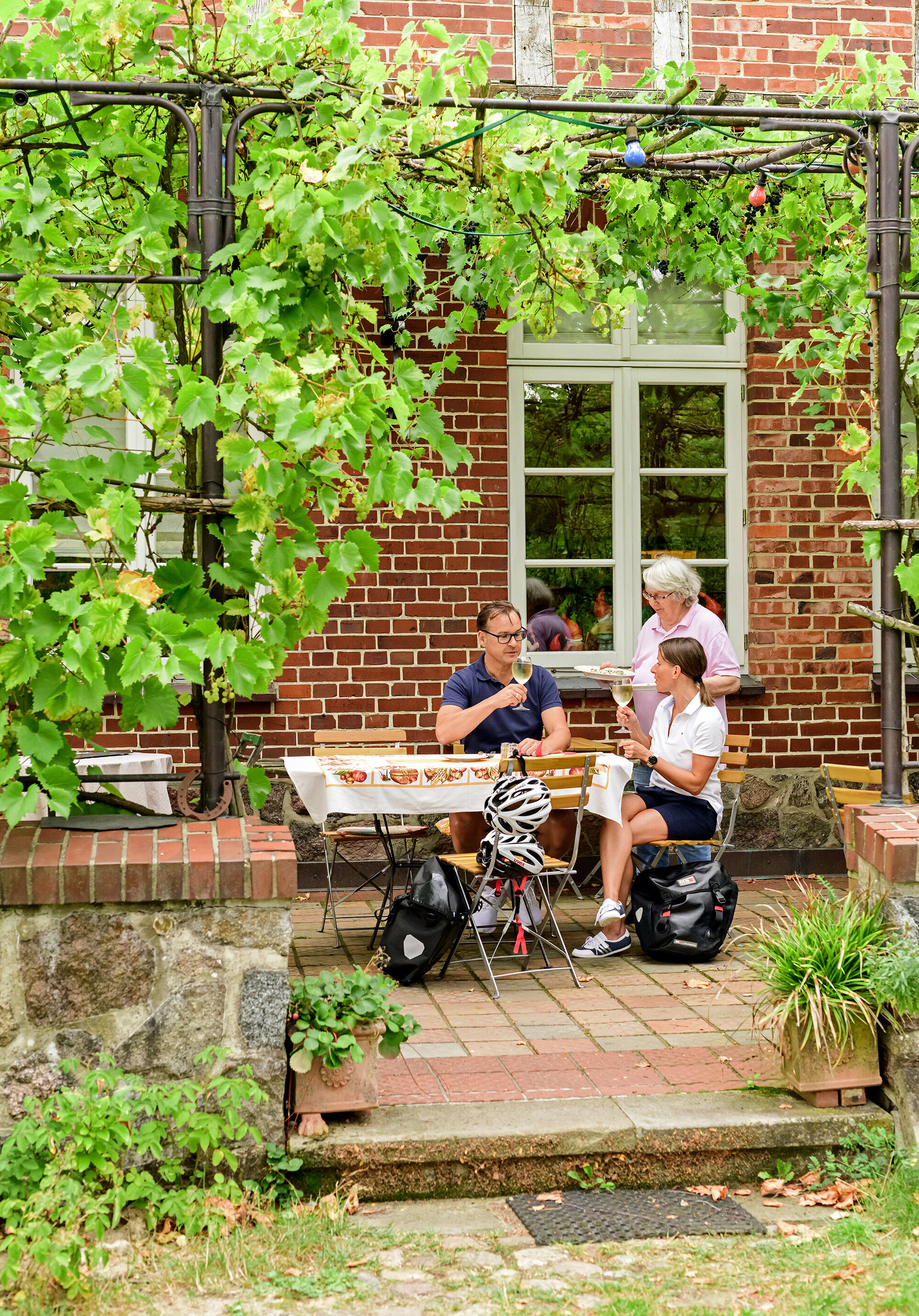 Ein Paar Radurlauber (Packtaschen und Fahrradhelme neben ihnen) sitzt auf der Terasse eines Restaurants und wird von der Wirtin bedient