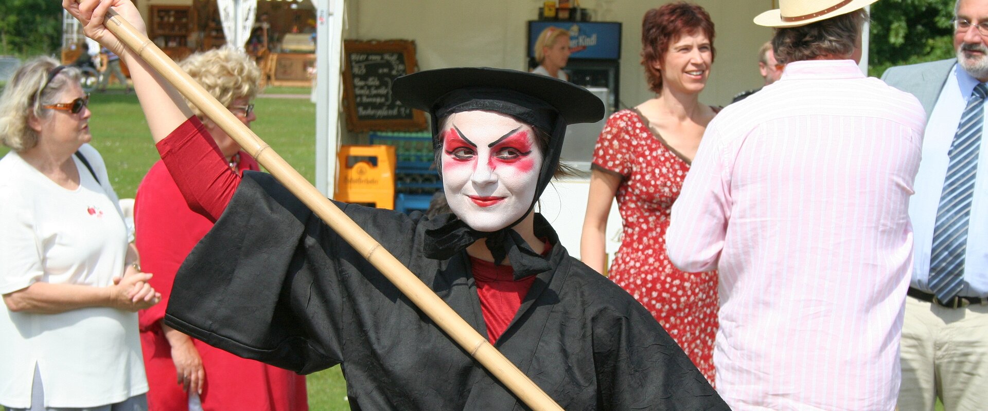 SchauspielerIN mit schwarzem Gewand, schwarzem Hut und weiß geschminktem Gesicht auf dem Kulturlustwandel in Bad Bevensen, im Hintergrund Festivalbesucher und einige Pagodenzelte