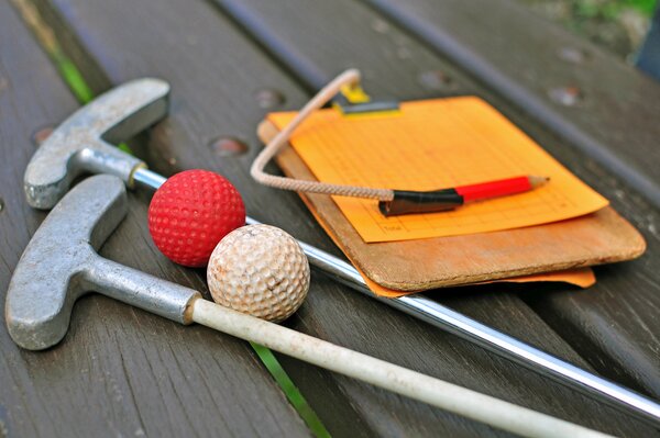 Zwei Minigolfschläger, ein roter und ein weißer Golfball und ein hölzernes Klemmbrett mit einem Spielplan und einem Stift liegen auf einer braunen Holzbank