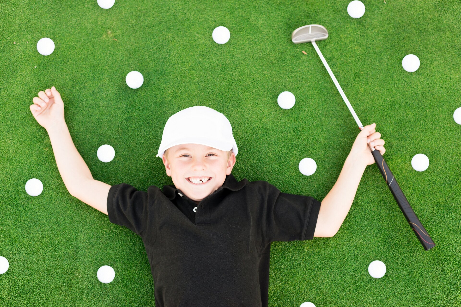 Der Oberkörper eines lachenden, zehnjährigern Jungen, bekleidet mit einem schwarzen T-Shirt und einer weißen Schirmmütze, liegt auf der Rasenfläche eines Golfplatzes, in der rechten Hand hält er einen Golfschläger, rings um ihn verteilt sind mehrere Golfbälle 