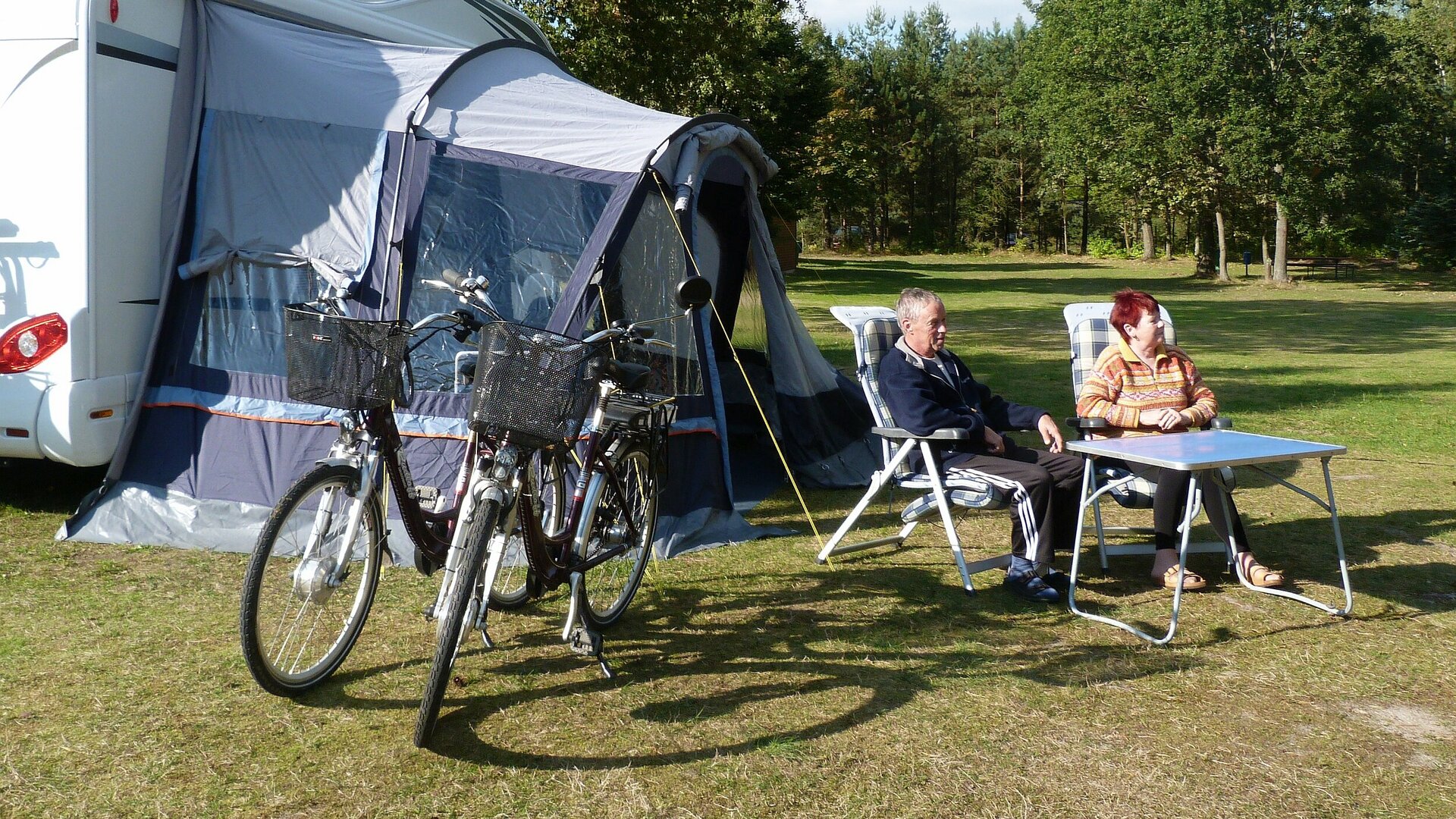 Ein Paar im Seniorenalter sitzt neben seinen Fahrrädern am Campingtisch vor einem Wohnmobil.