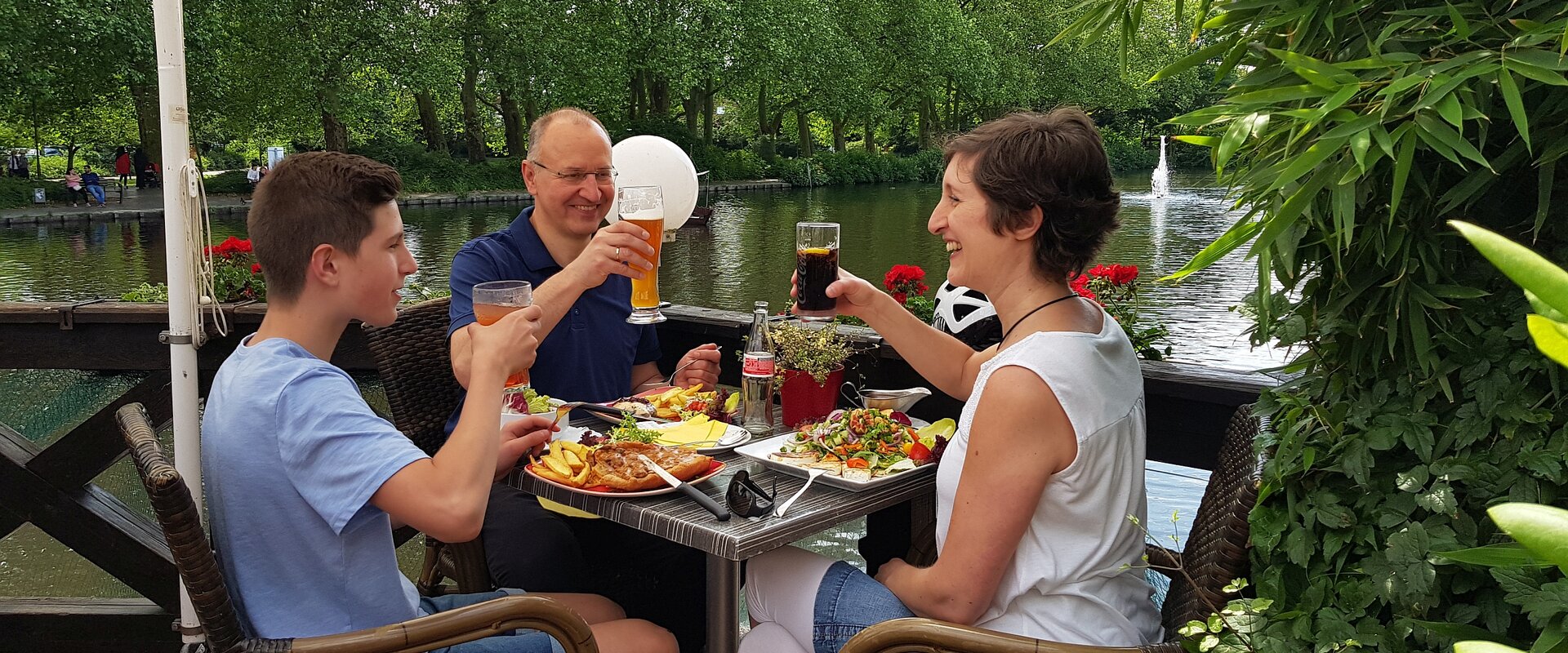 Eine sommerlich gekleidete Familie sitzt an einem mit Essen gedeckten Tisch auf einer Außenterasse eines Gastronomiebetriebes und prostet sich mit ihren Getränken zu