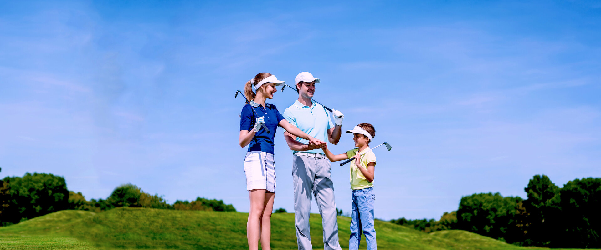 Eine junge Familie (Vater, Mutter und Sohn) stehen unter blauem Himmel in Golfkleidung auf dem Green eines Golfplatzes und haben idie Enden ihrer Schläger auf ihre Schultern gelegt