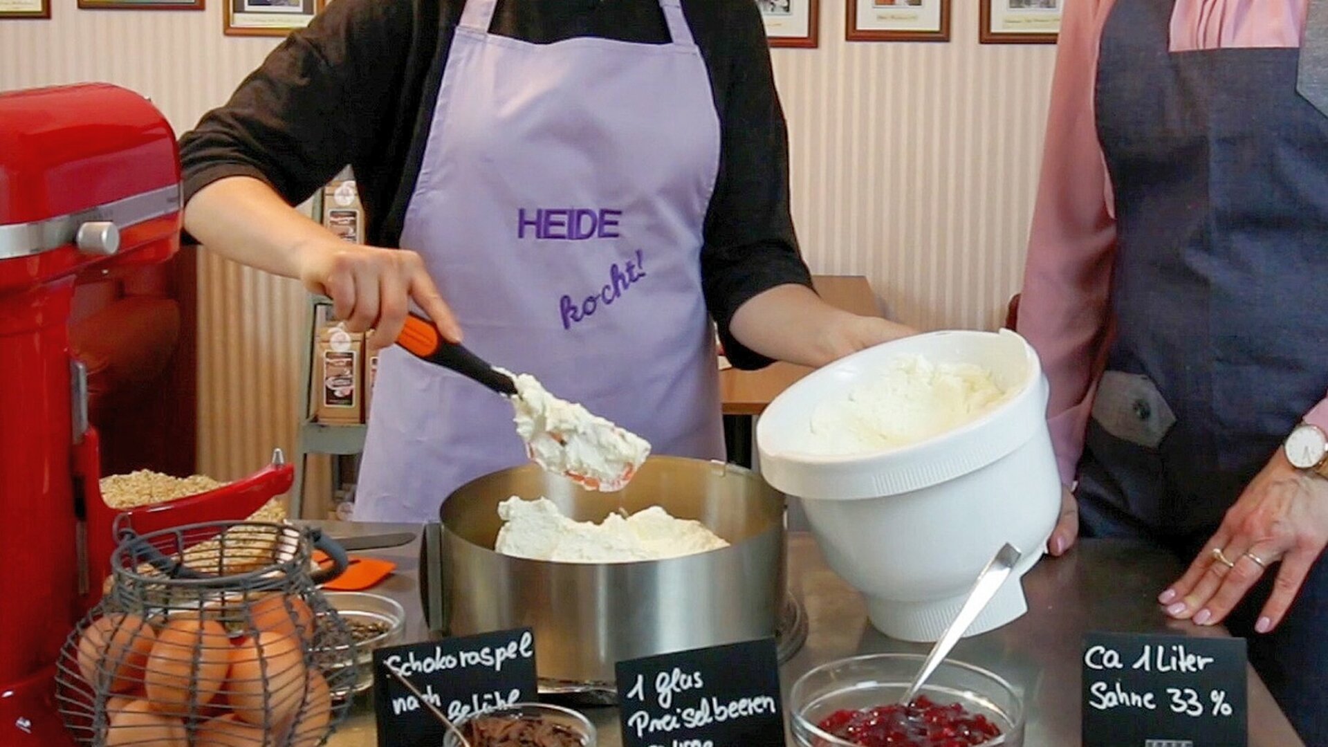 Zwei junge Frauen füllen geschlagene Buttercreme in eine runde Kuchenform. Die links hinter dem Tisch mit den Backzutaten stehende Frau trägt eine lila Schürze mit der Aufschrift "Heide kocht".