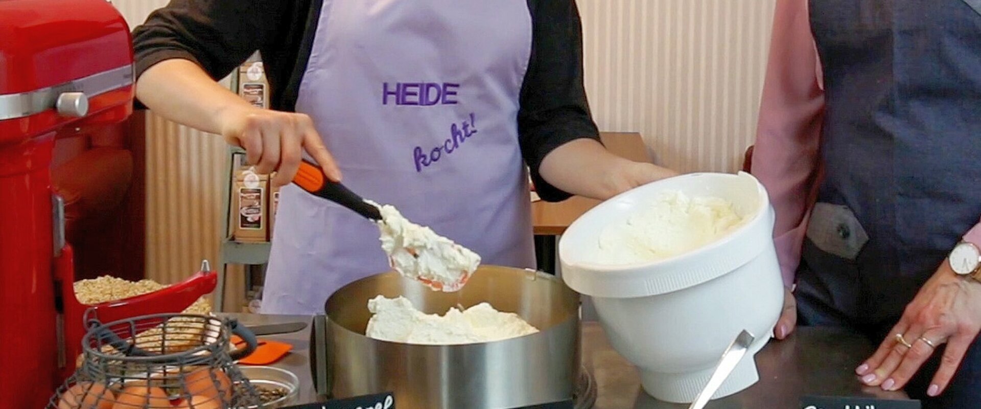 Zwei junge Frauen füllen geschlagene Buttercreme in eine runde Kuchenform. Die links hinter dem Tisch mit den Backzutaten stehende Frau trägt eine lila Schürze mit der Aufschrift "Heide kocht".